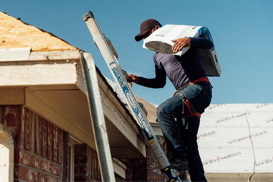 A roofer carries shingles up a ladder beside a home during roof replacement